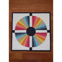 Draufsicht auf den fertigen Mini-Quilt mit vierteiligem Regenbogen-Kreismuster und dunkelblauem Kreuzrahmen auf Holzboden.